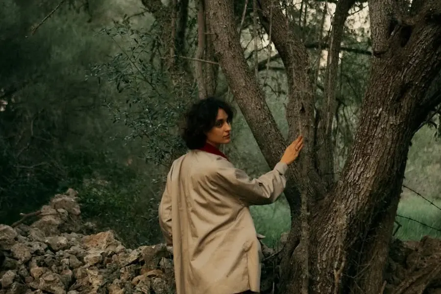 ema standing among the old trees on her land, one hand resting on a trunk
